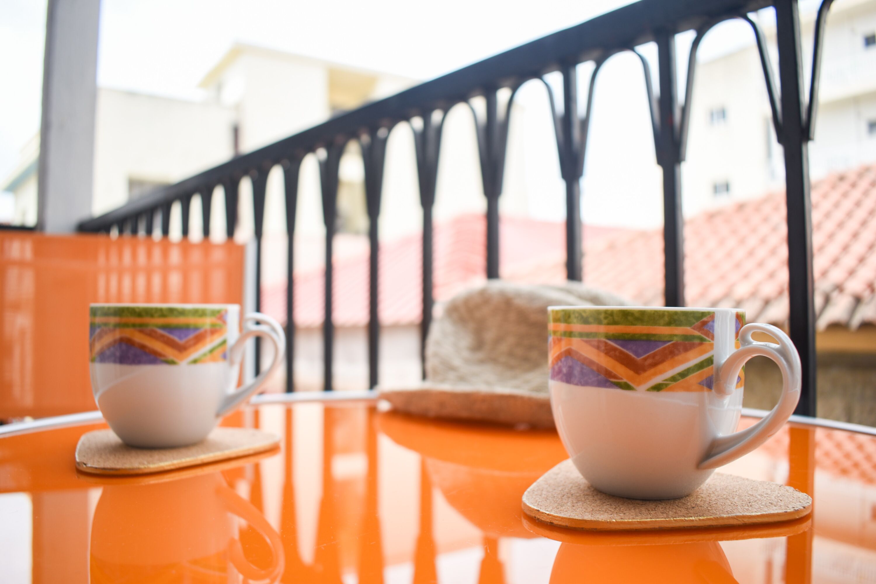 Two white cups with colorful patterns on a reflective orange table with coasters, set against a balcony railing with a view of buildings.