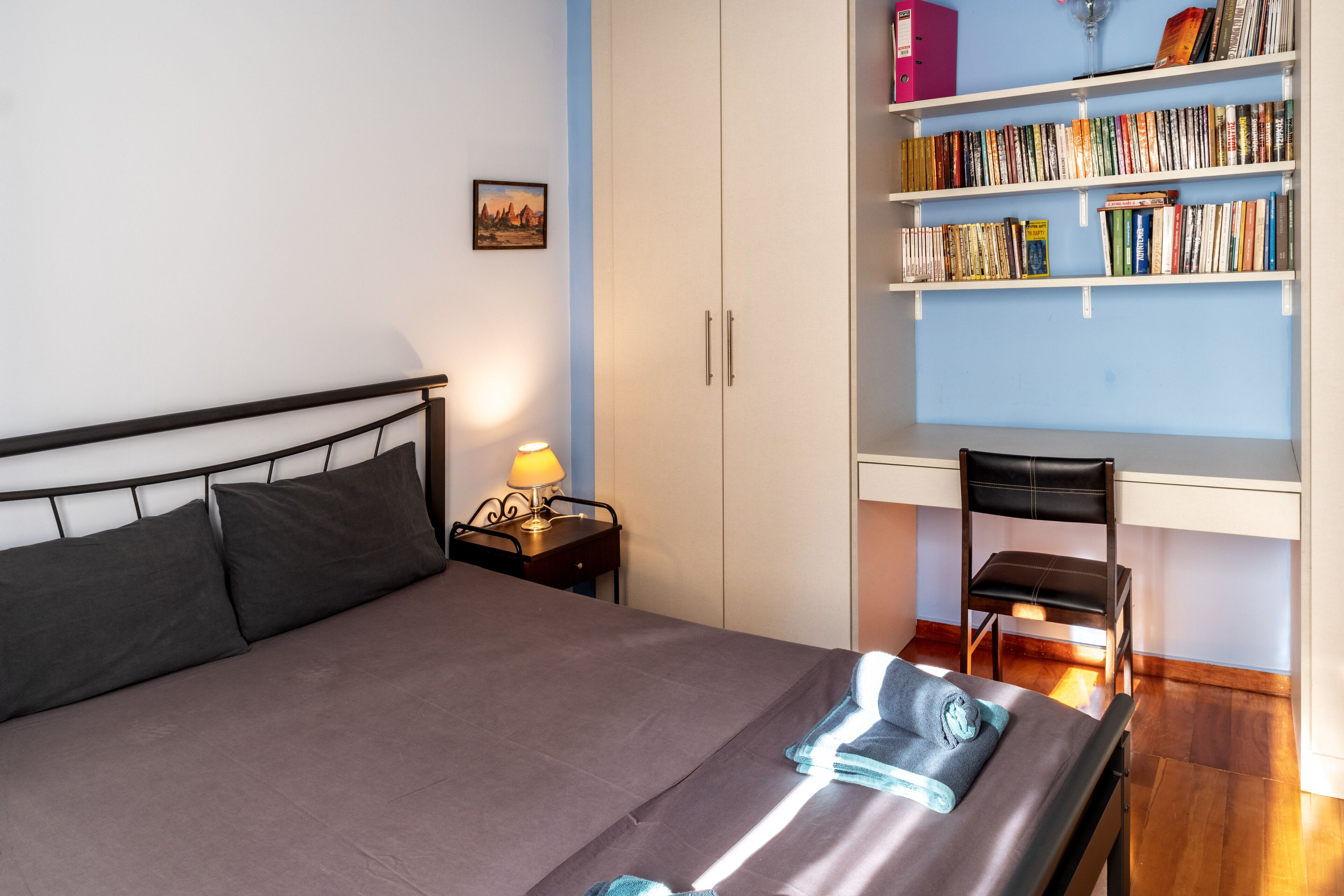 A bedroom with a bed, bedside table, lamp, bookshelf, and a desk with a chair.