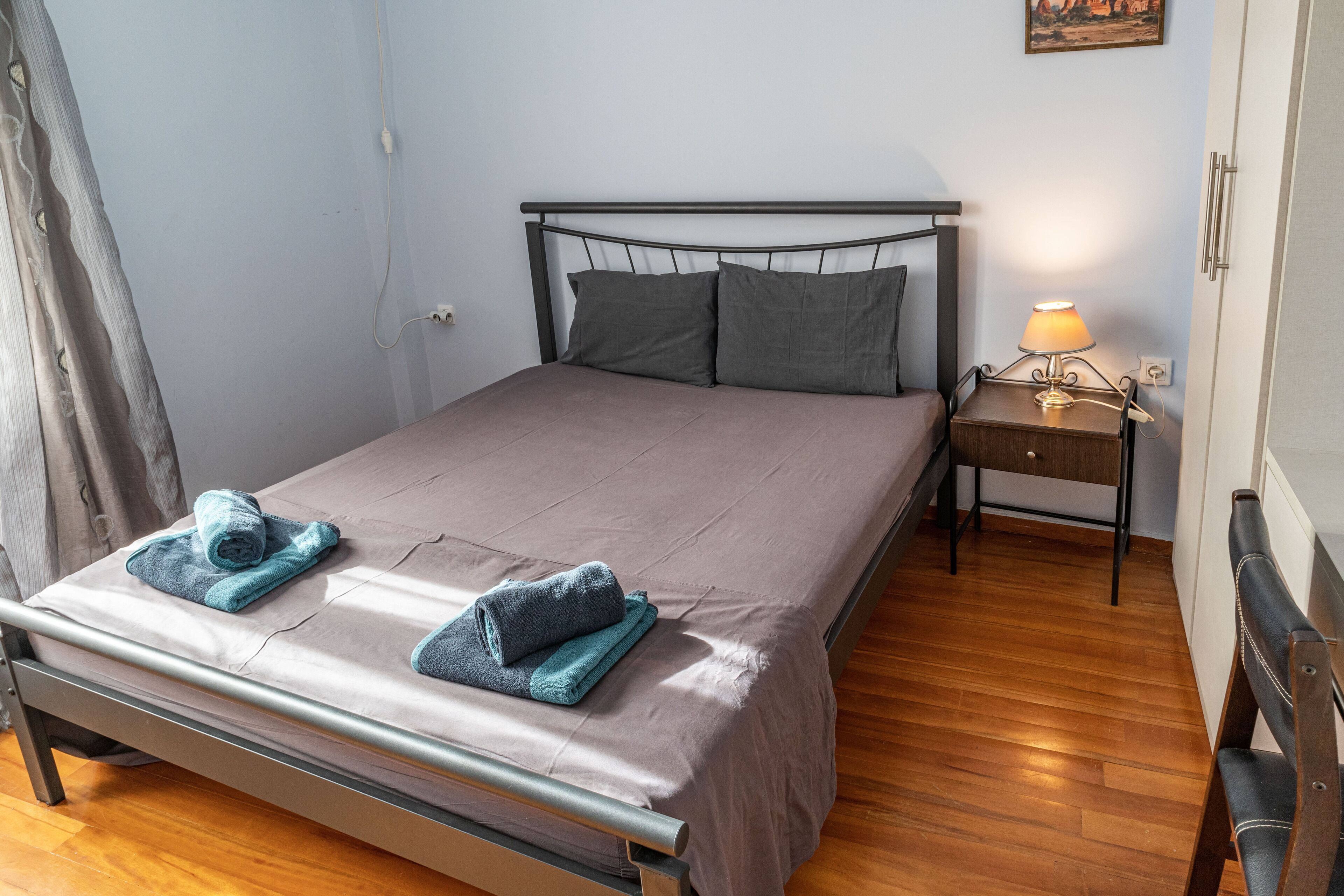 A neatly made bed with two pillows and two folded towels, a bedside table with a lamp, and a wooden floor.