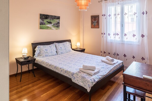 Un dormitorio con piso de madera, una cama con ropa de cama floral, dos mesitas de noche con lámparas, una ventana con cortinas florales y un cuadro en la pared.