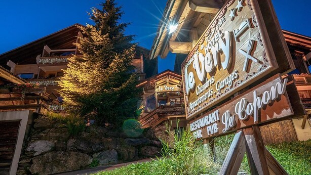 Uma placa de madeira indica um restaurante e um bar, emoldurada por uma paisagem montanhosa e uma grande árvore.