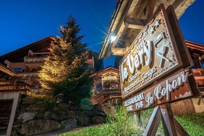 Une enseigne en bois portant l’inscription d’un restaurant et d’un bar, le tout situé dans un paysage montagneux et bordé d’un grand arbre.