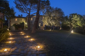 A stone pathway leading to a stone building with lit windows and trees. - Casale degli Ulivi (Rapolano Terme)