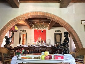 A dining area with a brick archway, wooden beams, and a chandelier. There are tables set with plates, fruits, and a vase of flowers. - Casale degli Ulivi (Rapolano Terme)