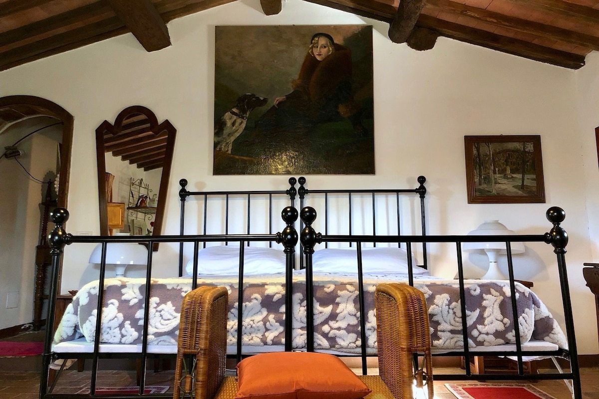A bedroom with a bed, two wicker chairs, a painting, and a mirror.