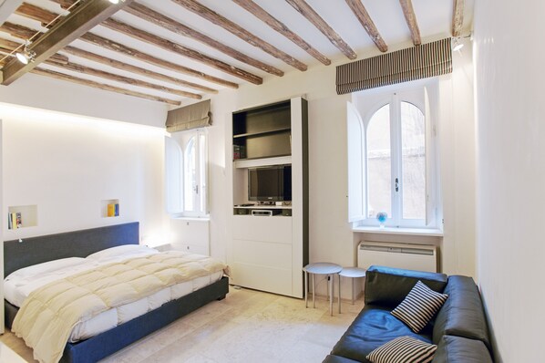 A bedroom with a bed, a built-in TV, a small table, a sofa, and a large window. - Oro - WR Apartments near Castel Sant'Angelo (Rome)