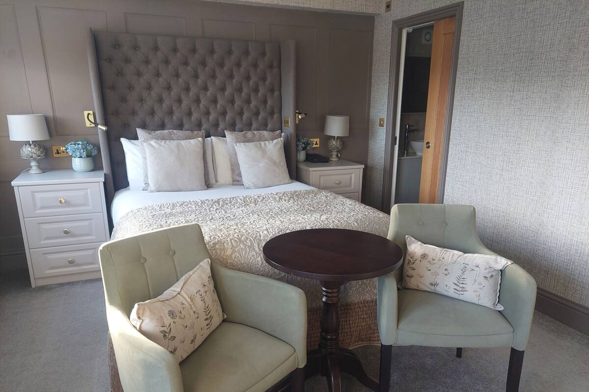 A hotel room with a large bed, two armchairs, a small round table, and a nightstand with lamps.
