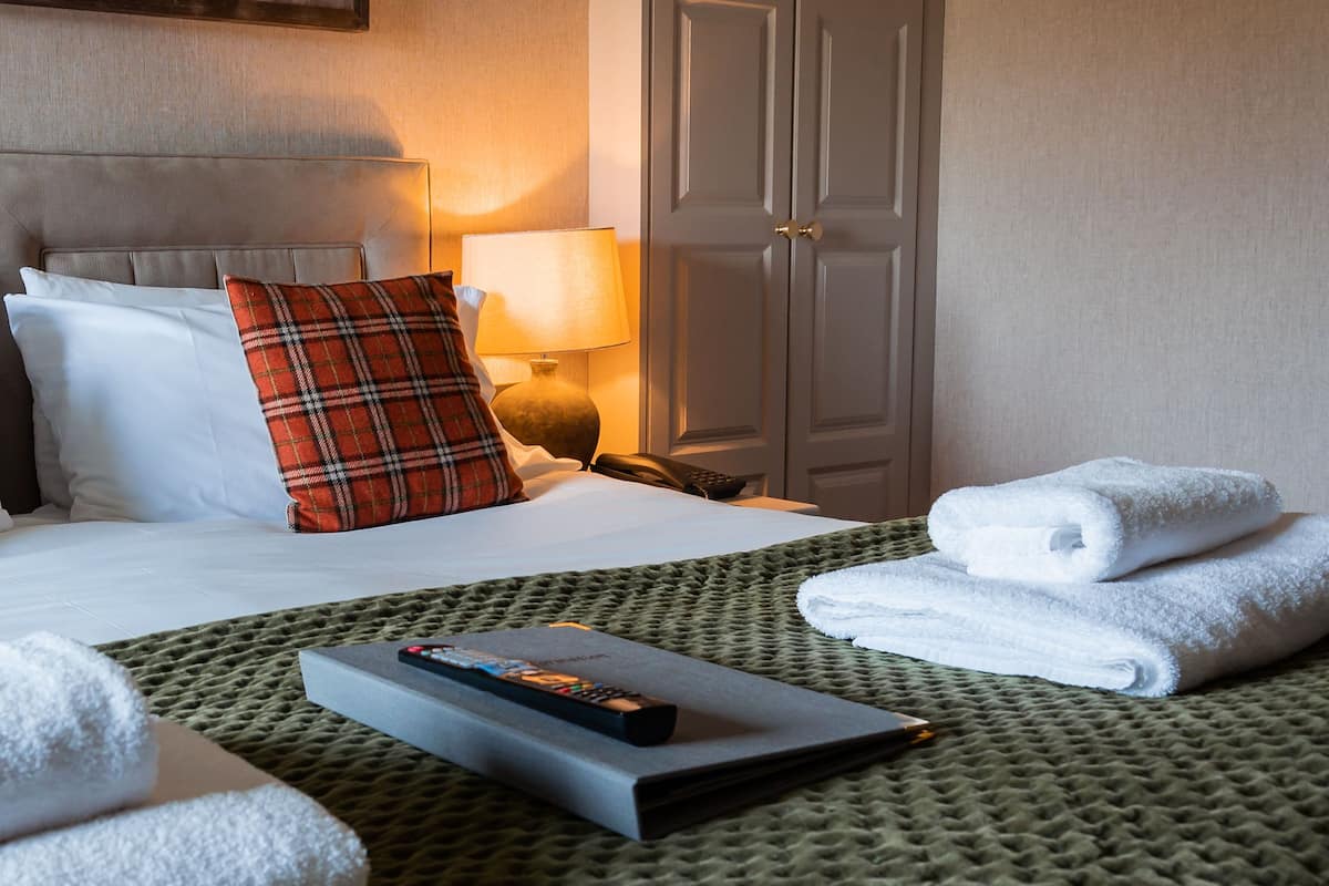 A neatly made bed with white and plaid pillows, a remote control, and folded towels on a green textured bedspread.
