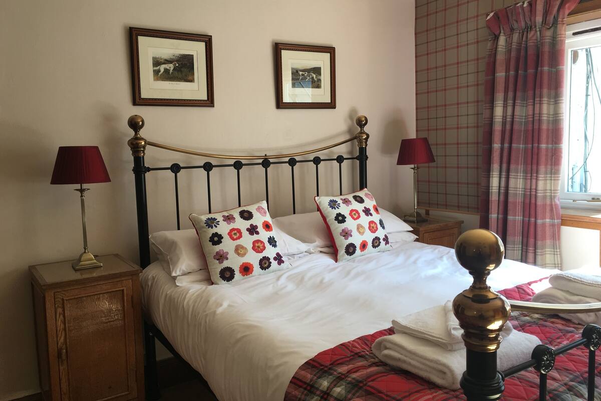 A neatly made bed with floral pillows, a plaid blanket, and a gold headboard. There are two framed pictures on the wall above the bed.