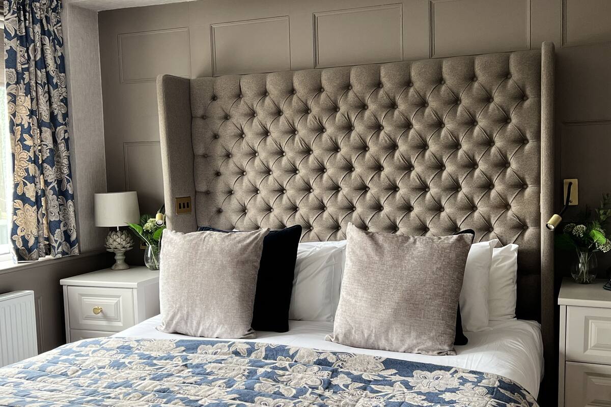 A bedroom with a tufted headboard, a floral-patterned bedspread, and a nightstand with a lamp and a vase of flowers.
