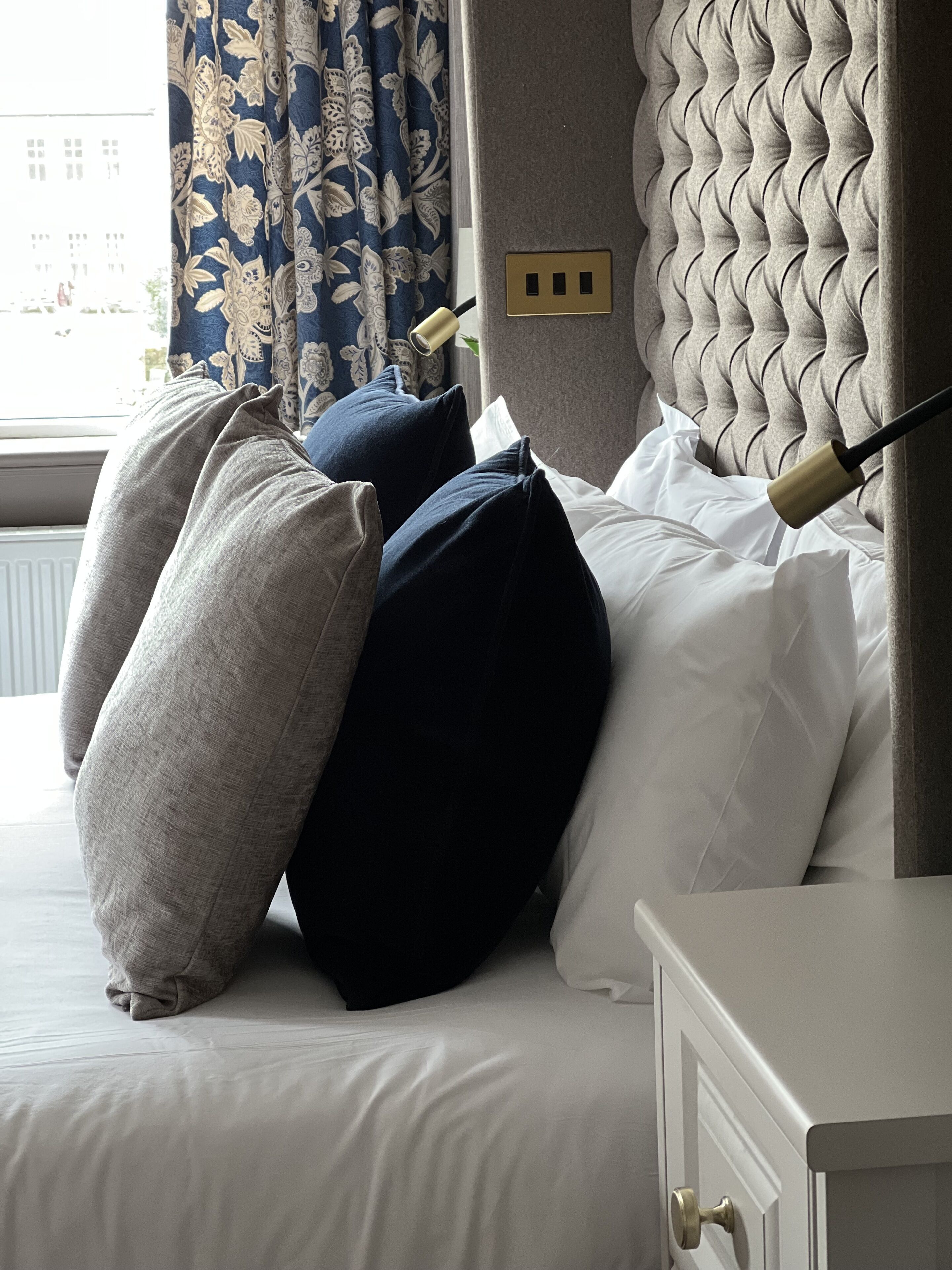 Un letto rifatto con cura, con testiera trapuntata, una finestra con tende e un comodino con una lampada.