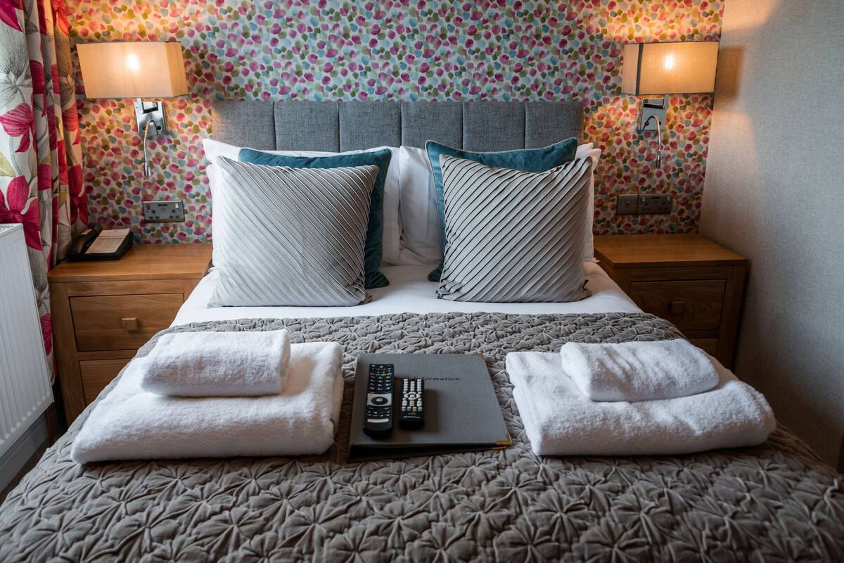 A neatly made bed with pillows, a remote control, and towels on a patterned bedspread.