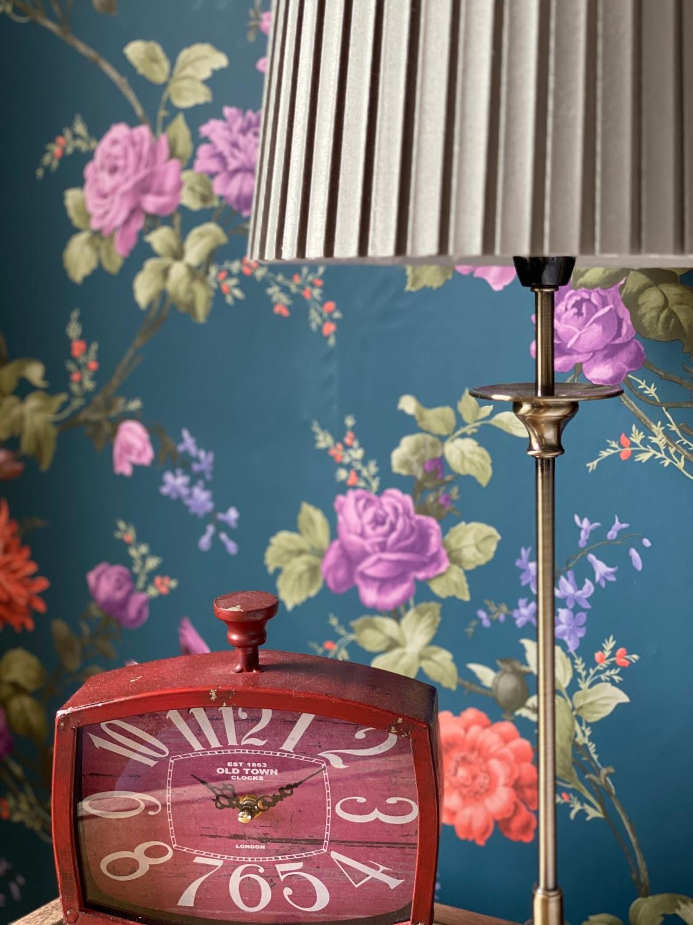 A vintage red clock with a brass stand, set against a floral wallpaper background.