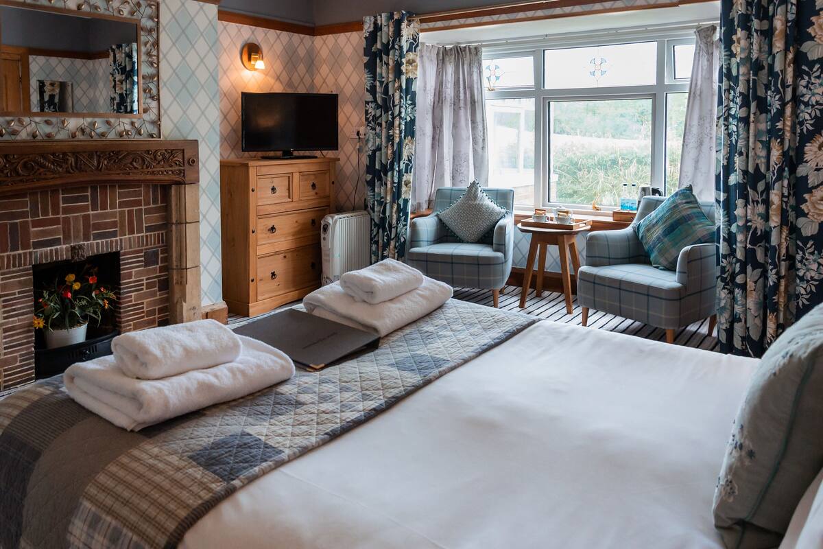 A well-appointed hotel room with a large bed, a fireplace, a television, and a cozy seating area by the window.