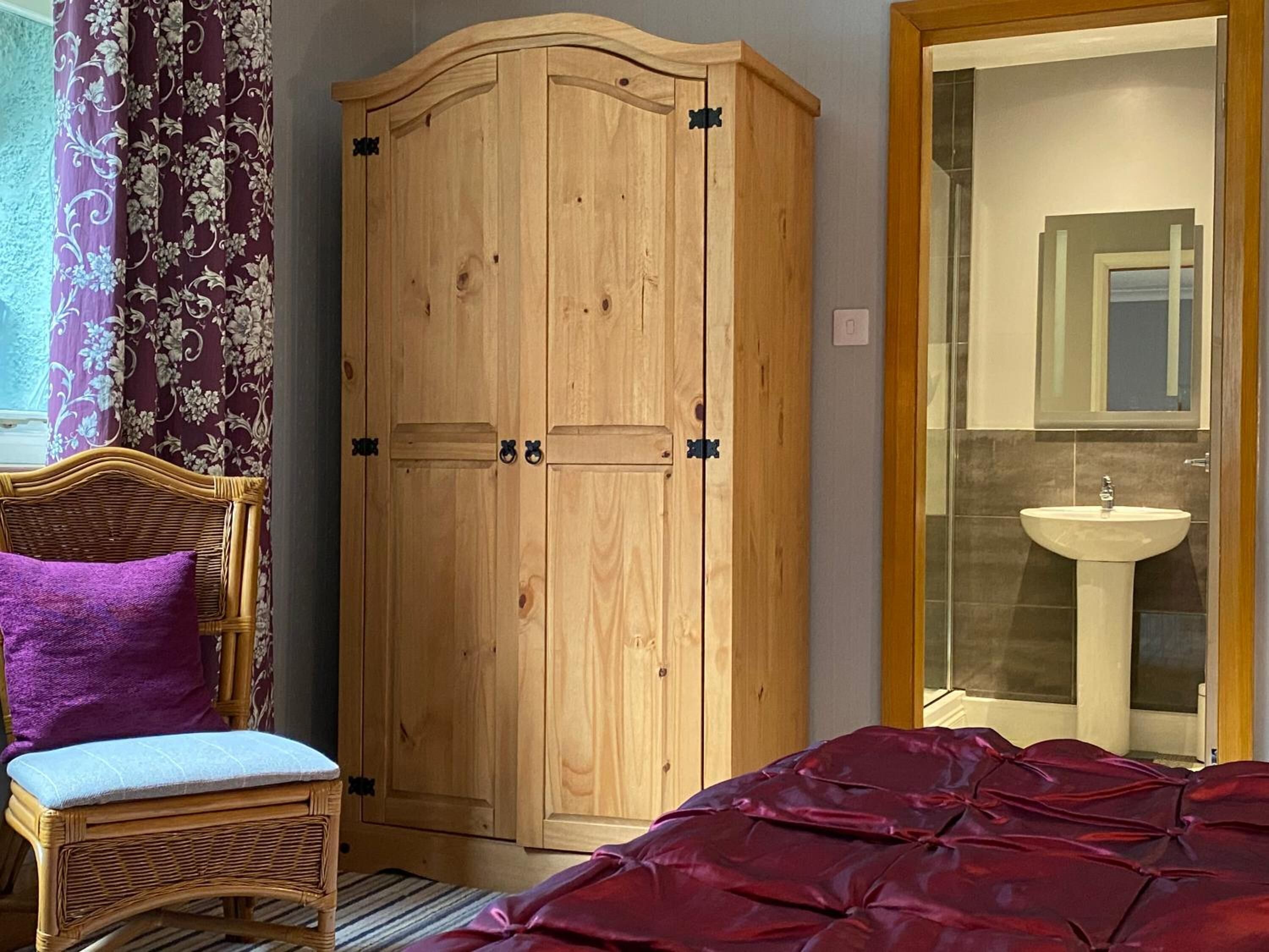 A bedroom with a wooden wardrobe, a wicker chair with a purple cushion, a red bedspread, and a bathroom with a sink and mirror.