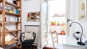 A cozy room with a wooden bookshelf, a white desk with a lamp, a chair, and a view of a balcony with potted plants. - Otranto - WR Apartments (Rome)