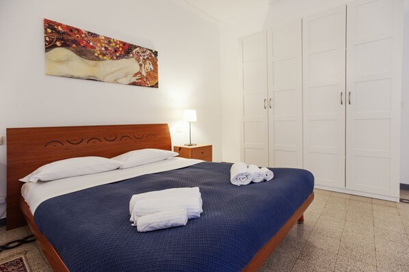 A neatly made bed with a blue bedspread and white pillows, a wooden headboard, a bedside lamp, and a painting on the wall. - Palestrina - WR Apartments (Rome)