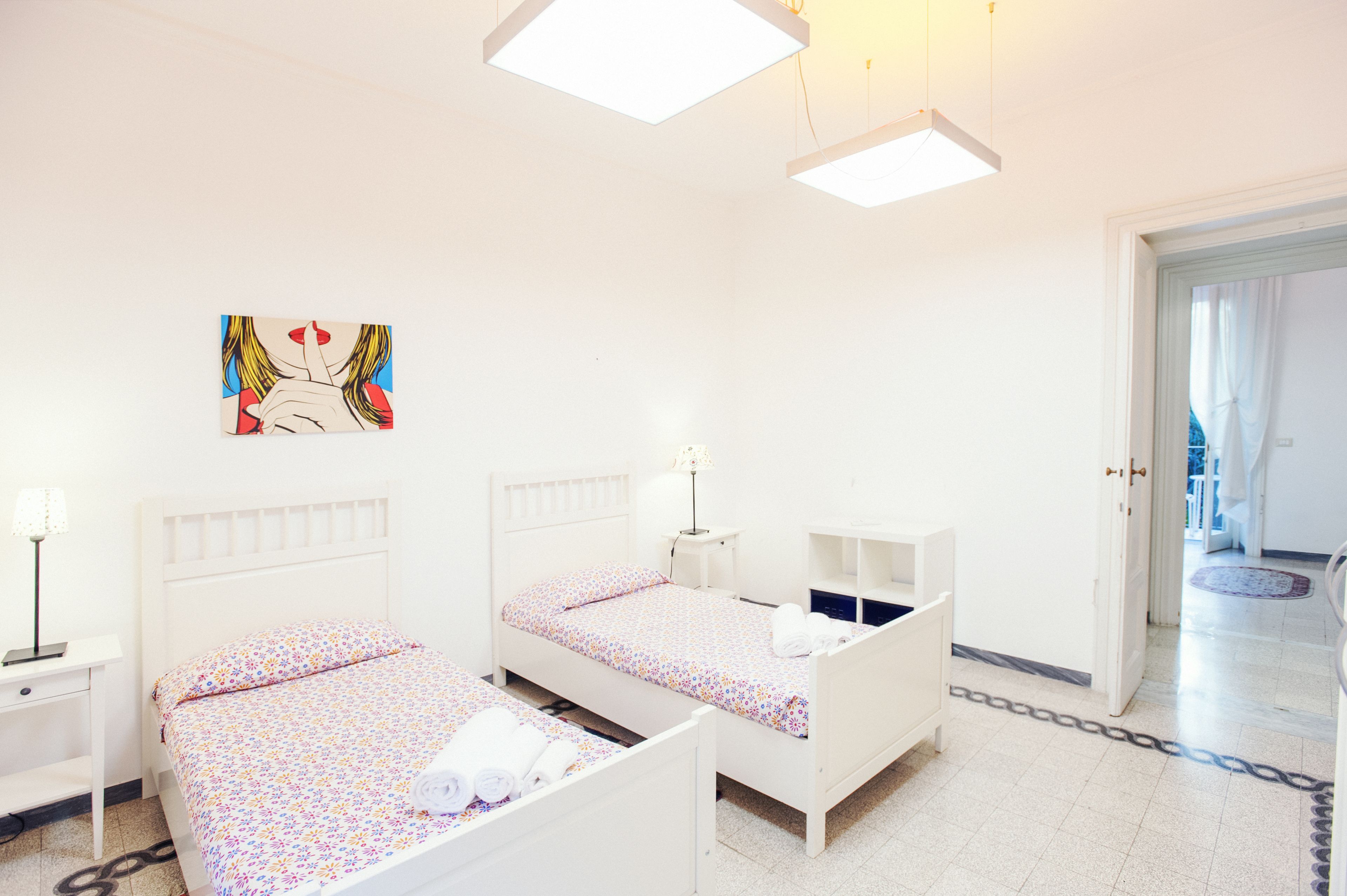 A hotel room with two single beds, a small bedside table with a lamp, a wardrobe, and a painting on the wall.