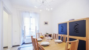 A dining area with a wooden table set for four, surrounded by chairs. There is a window with sheer curtains, a small table with a bowl and fruit, and a wall with decorative panels. - Palestrina - WR Apartments (Rome)