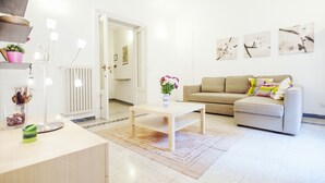 A modern living room with a beige sofa, a coffee table, and a vase of flowers. There is a white door leading to another room, and a radiator on the wall. - Palestrina - WR Apartments (Rome)