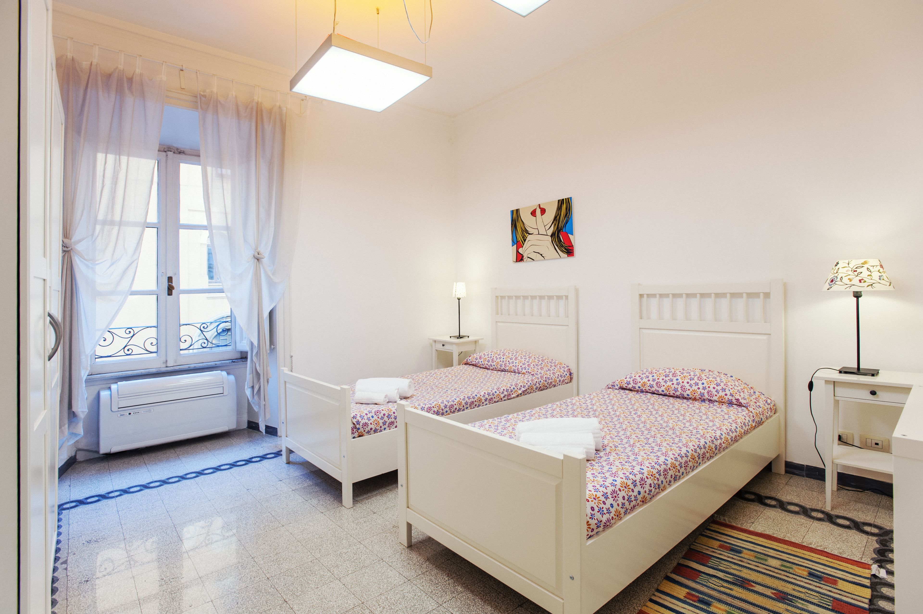 A room with two beds, a radiator, a window with curtains, a painting, a lamp, and a patterned rug.