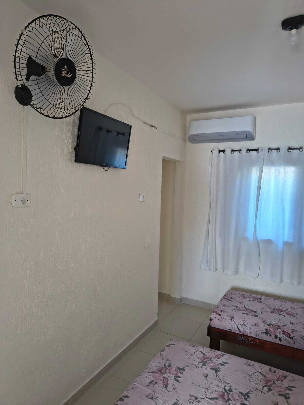 Una habitación con ventilador de pared, televisión, equipo de aire acondicionado, cama con colcha floral y ventana con cortinas.