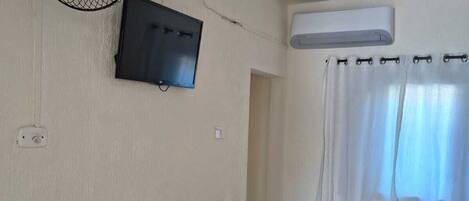 Una habitación con ventilador de pared, televisión, equipo de aire acondicionado, cama con colcha floral y ventana con cortinas.