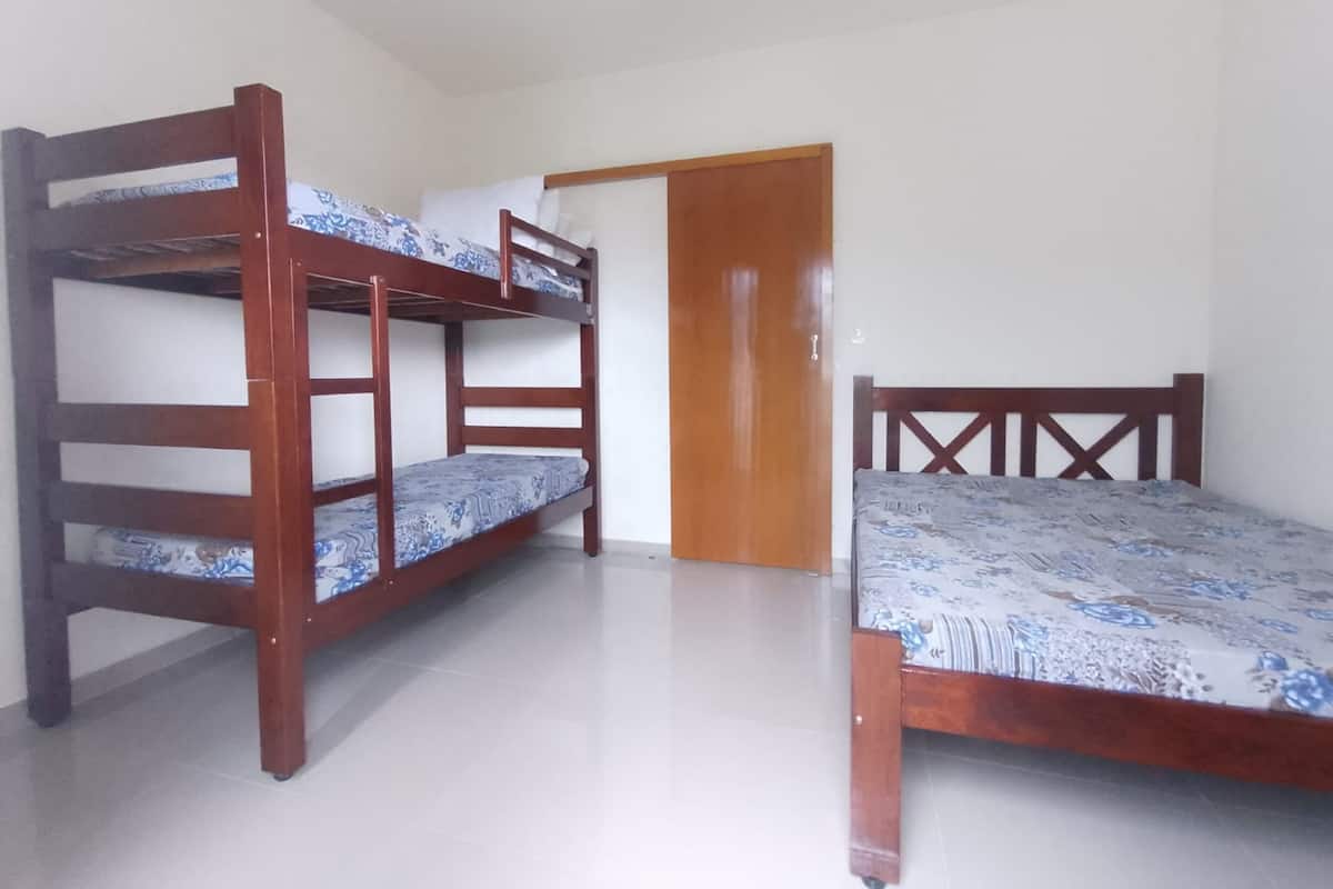 Quarto com duas beliches e uma cama de solteiro, porta de madeira e piso ladrilhado.