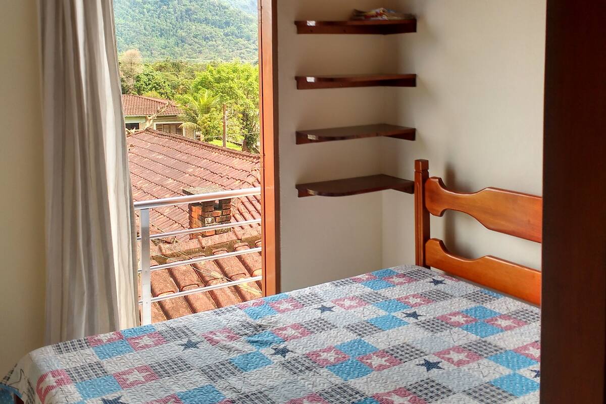 Quarto com teto de madeira, leito com colcha, janela com cortinas e vista para uma paisagem montanhosa.