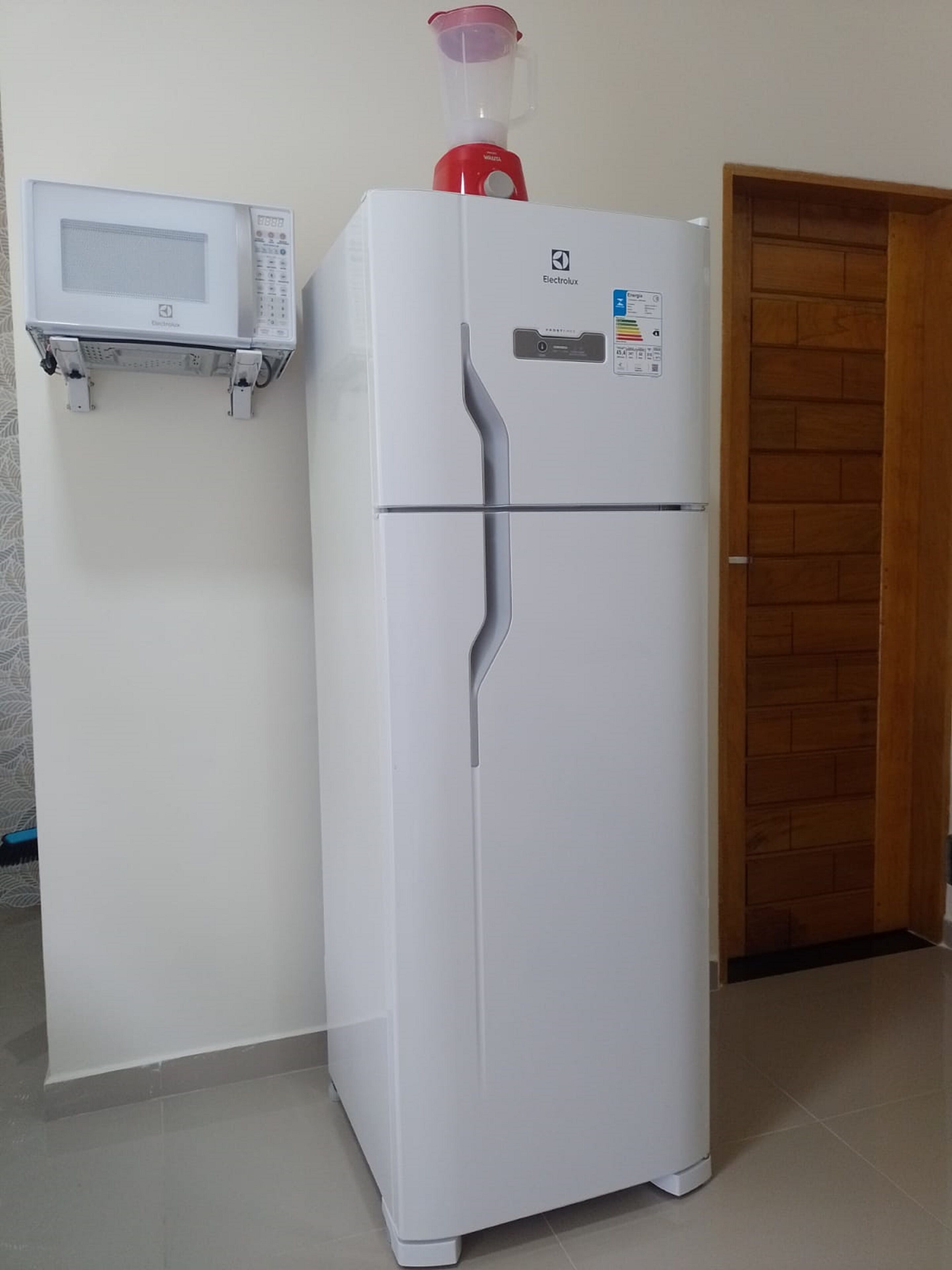 A white refrigerator with a red blender on top, a microwave on a shelf above it, and a wooden door in the background.