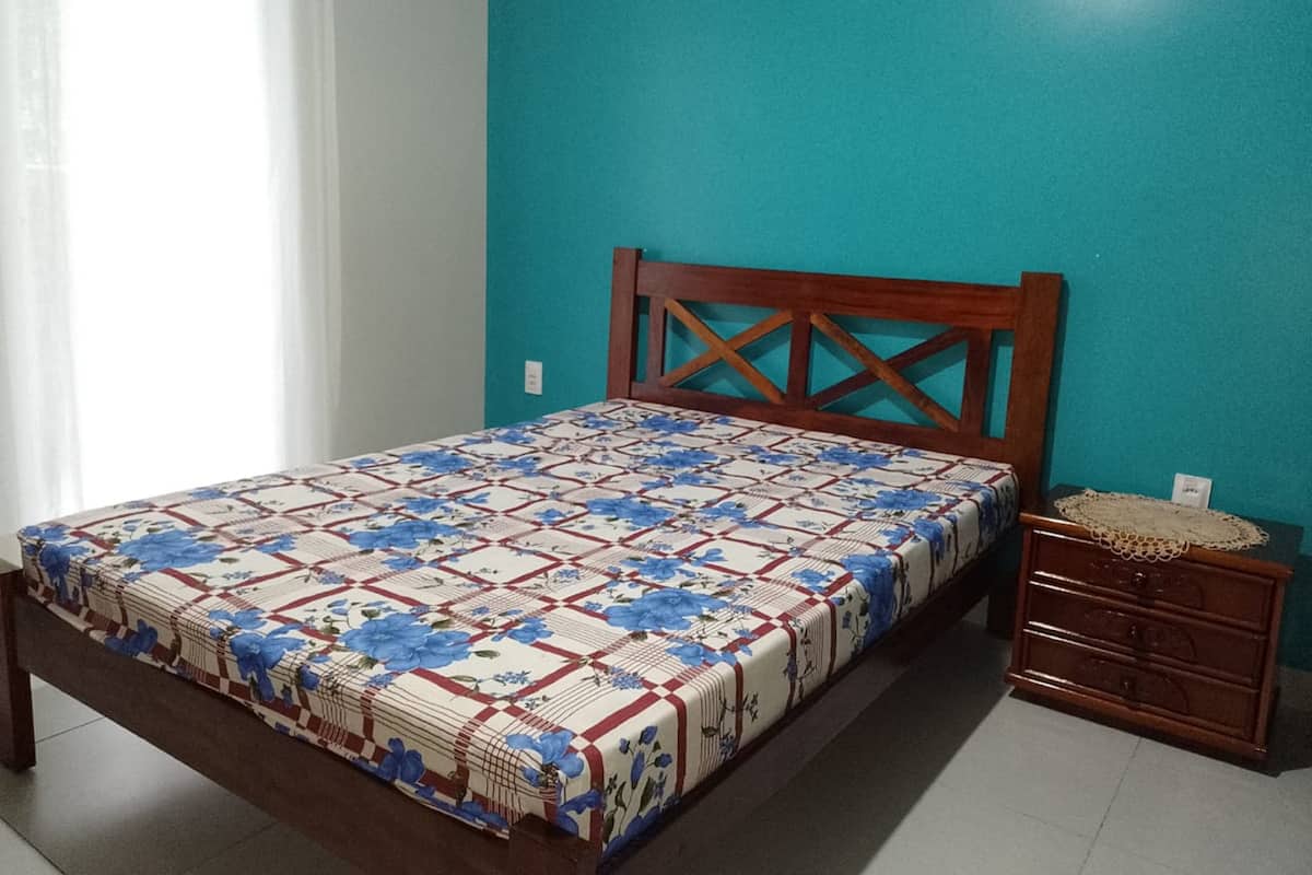 Quarto com cama, mesa de cabeceira de madeira e parede azul-esverdeada.