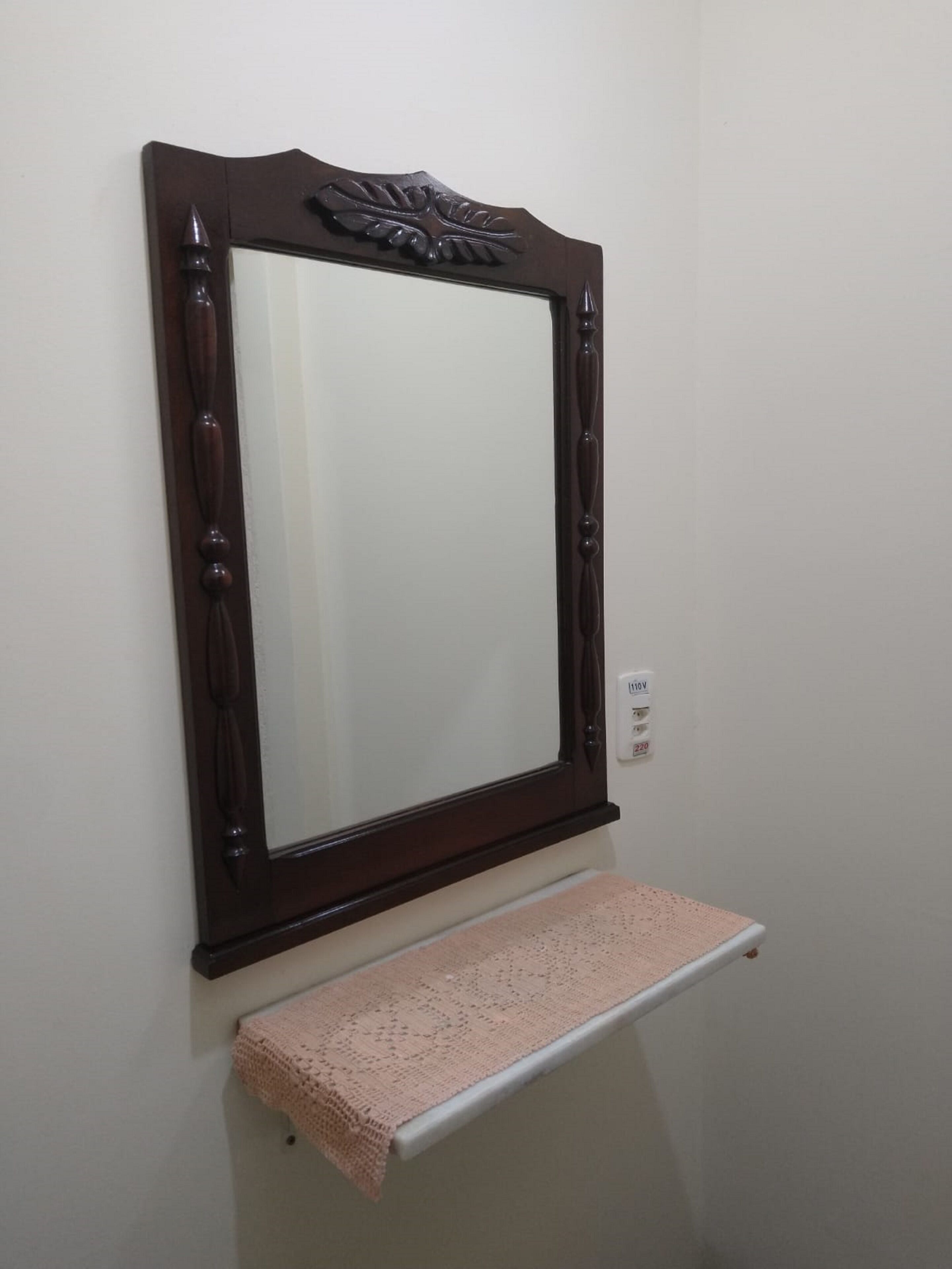 A mirror with a dark wooden frame and a decorative leaf motif at the top. Below the mirror, there is a white shelf with a pink crocheted item on it.