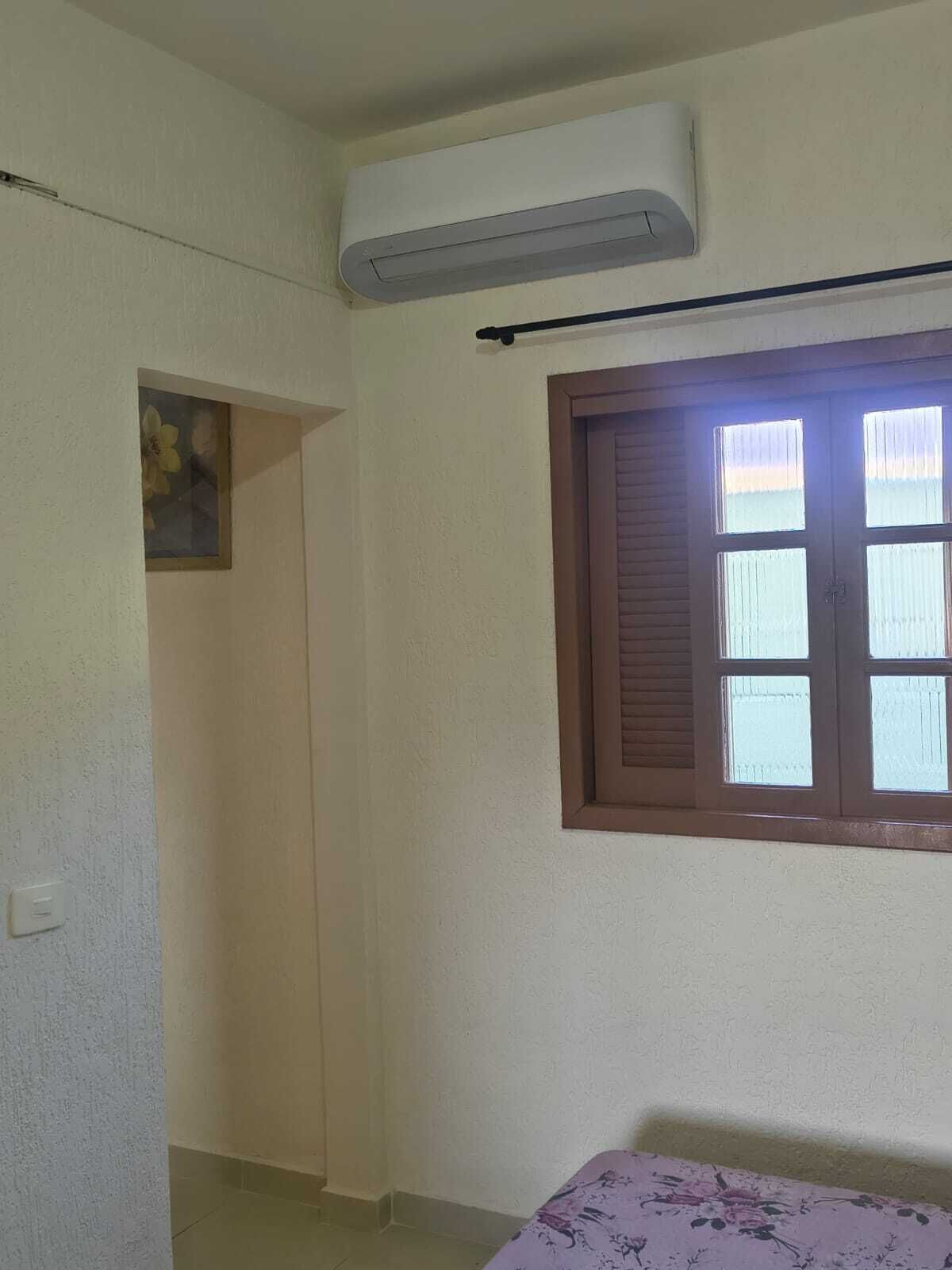 A room with a bed, an air conditioning unit, a window with blinds, and a small painting on the wall.