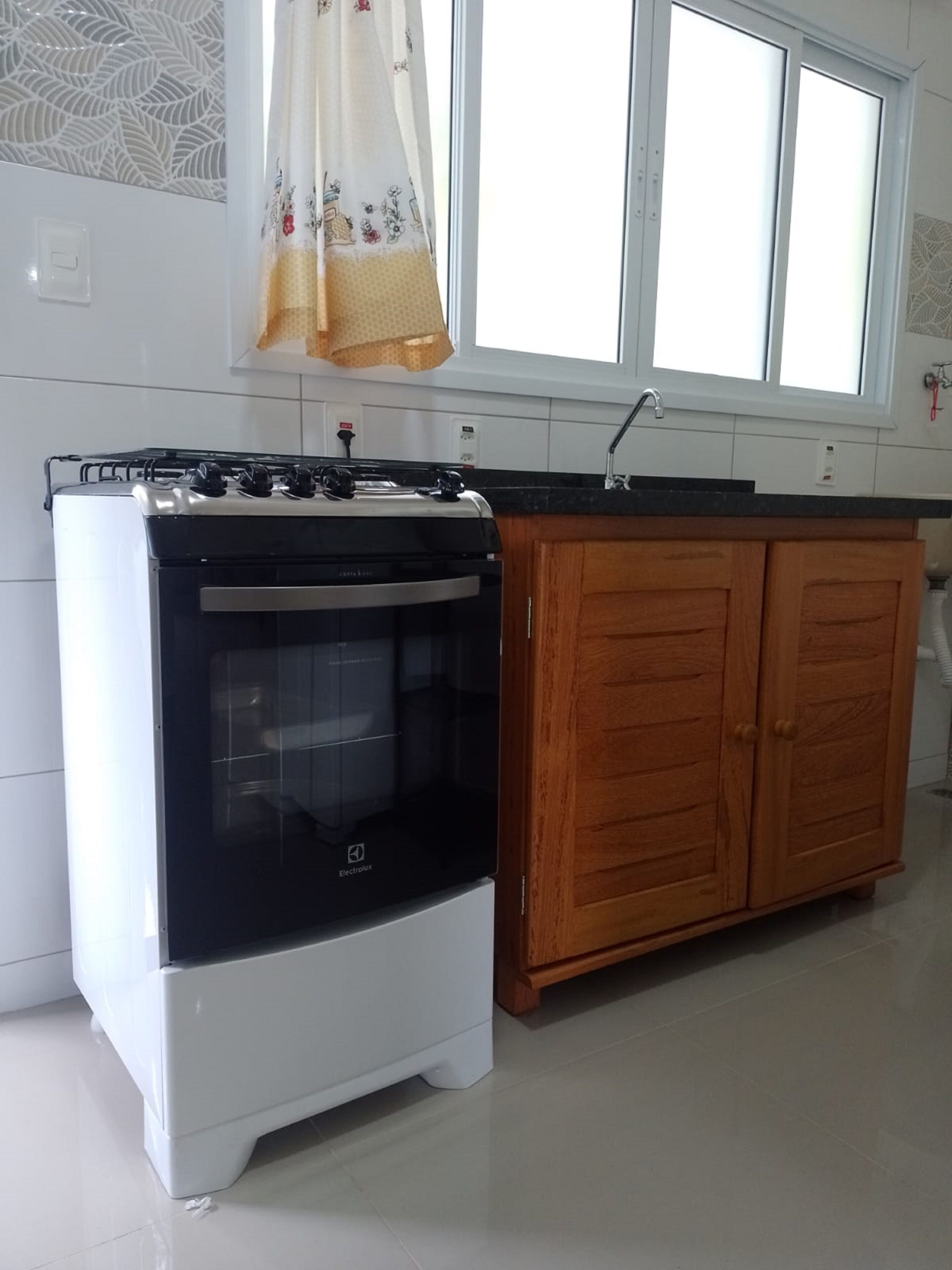 A kitchen with a white and black stove, wooden cabinets, a window with a curtain, and a tiled backsplash.