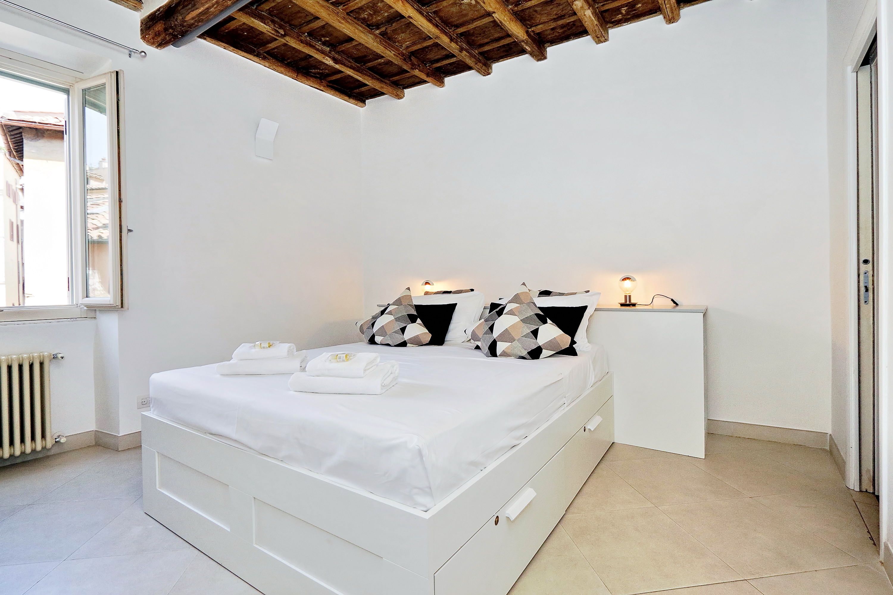 A bedroom with a white bed, a wooden ceiling, a bedside table with a lamp, and a window with a view of buildings.