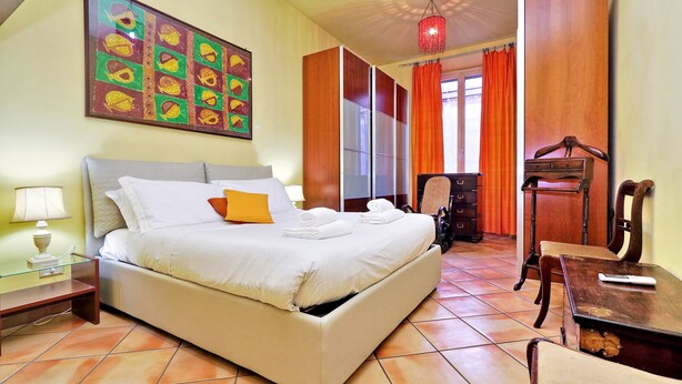 A hotel room with a bed, a desk, a chair, a lamp, and a painting on the wall. - Romalibera - WR Apartments (Rome)