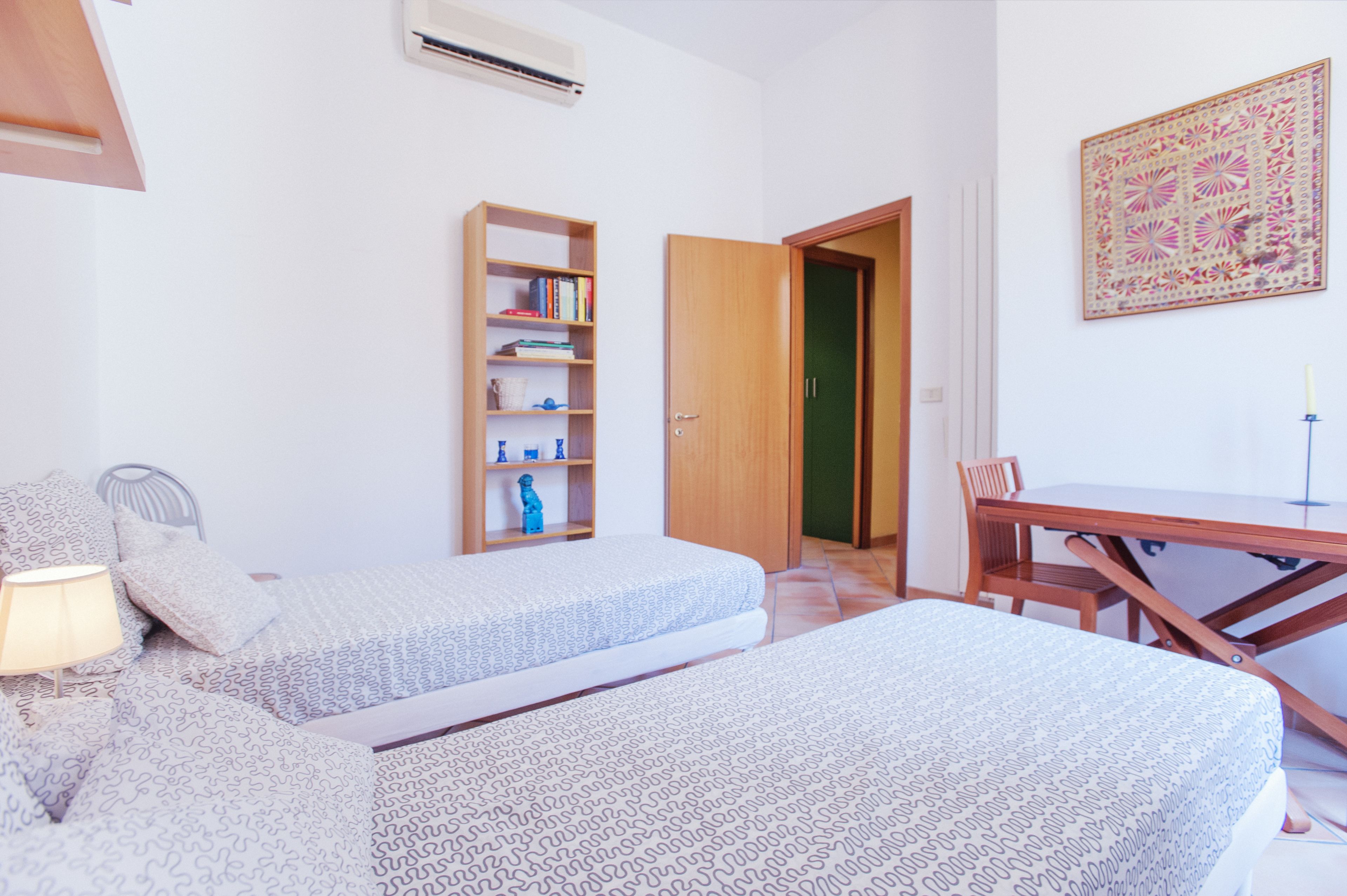 A room with two beds, a wooden desk, a chair, a bookshelf, and a door leading to another room.