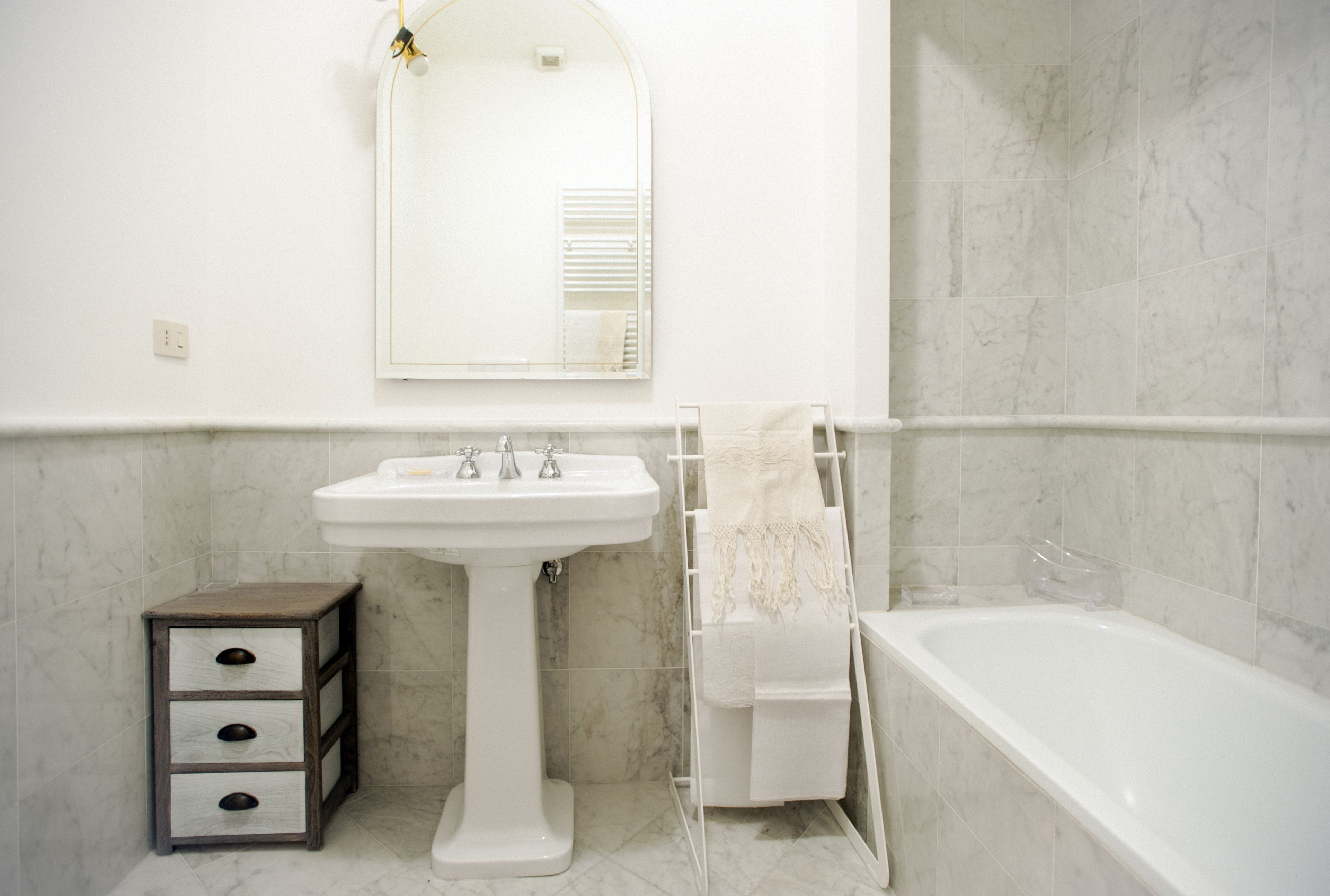 A bathroom with a pedestal sink, a mirror, a towel rack, a small wooden cabinet, and a bathtub.