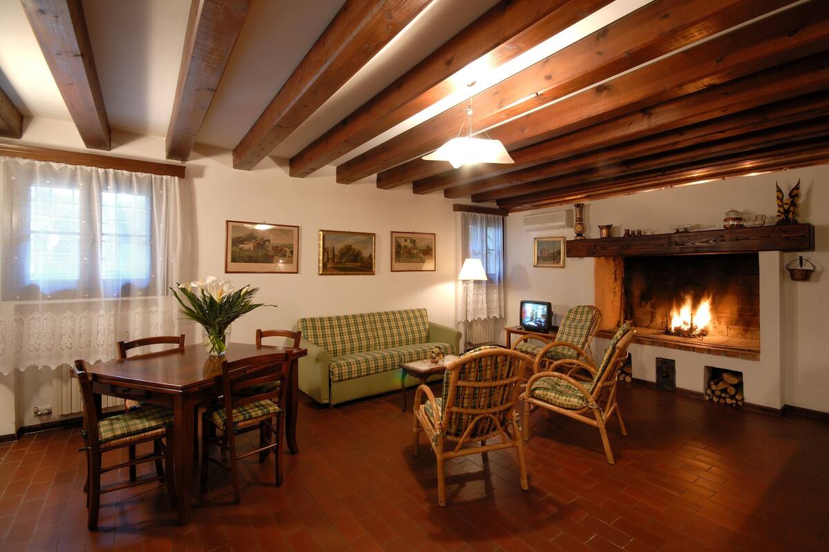 A cozy living room with a fireplace, wooden beams, a green sofa, and a dining table with chairs.
