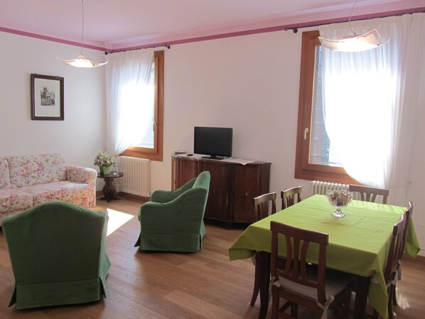A living room with a sofa, two green chairs, a dining table with a green tablecloth, a television on a wooden cabinet, and a framed picture on the wall. - Agriturismo Villa Selvatico (Vigonza)