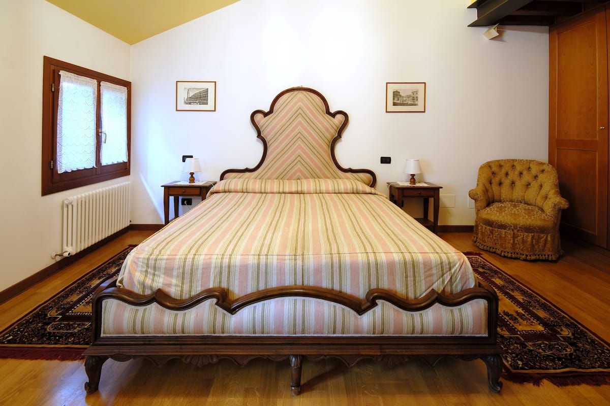 A bedroom with a large bed, wooden floor, two bedside tables, a chair, and a radiator.