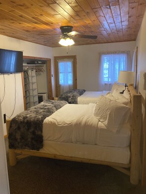 Un dormitorio con techo de madera, una cama con ropa de cama blanca, un ventilador de techo, un televisor montado en la pared y una ventana con cortinas traslúcidas.