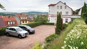 Zona residencial con una casa grande y blanca, dos coches aparcados y vistas a un paisaje urbano lejano.