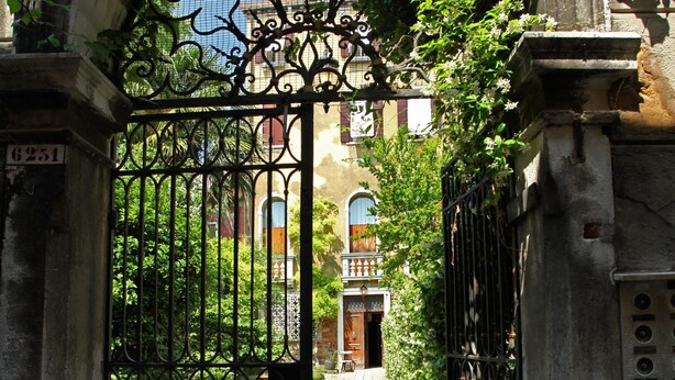 A wrought iron gate with a decorative arch, leading to a building with a brick facade and arched windows. - Testa - WR Apartments (Venice)