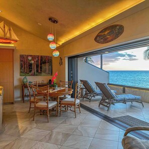 A dining area with a round table and chairs, a view of the sea, and hanging decorative lights.