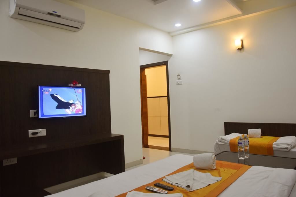 A hotel room with a TV mounted on the wall, a bed with a wooden headboard, a bedside table with a remote control, and a water bottle on the bed.
