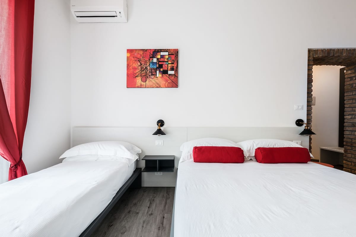A hotel room with two beds, a red curtain, a modern painting, a brick wall, and a small table with a lamp.
