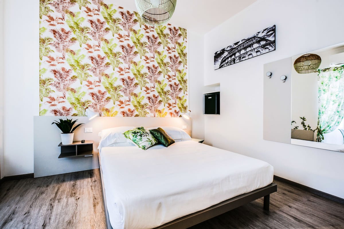 A modern bedroom with a large bed, a wall-mounted shelf with a plant, a mirror, and a patterned wallpaper.