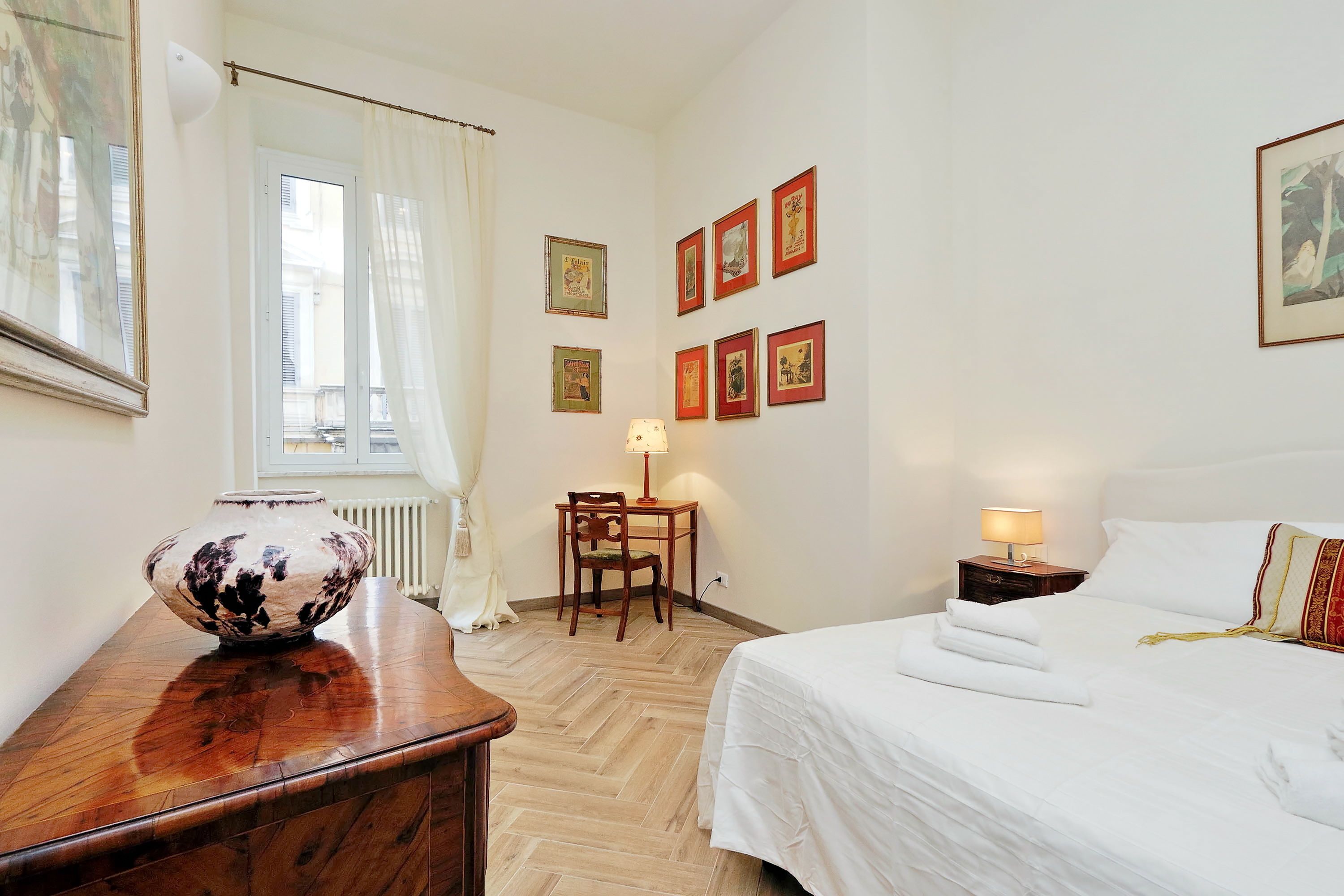Quarto de hotel com piso de madeira, cama com roupa de cama branca, escrivaninha com cadeira, abajur e várias fotos emolduradas na parede.