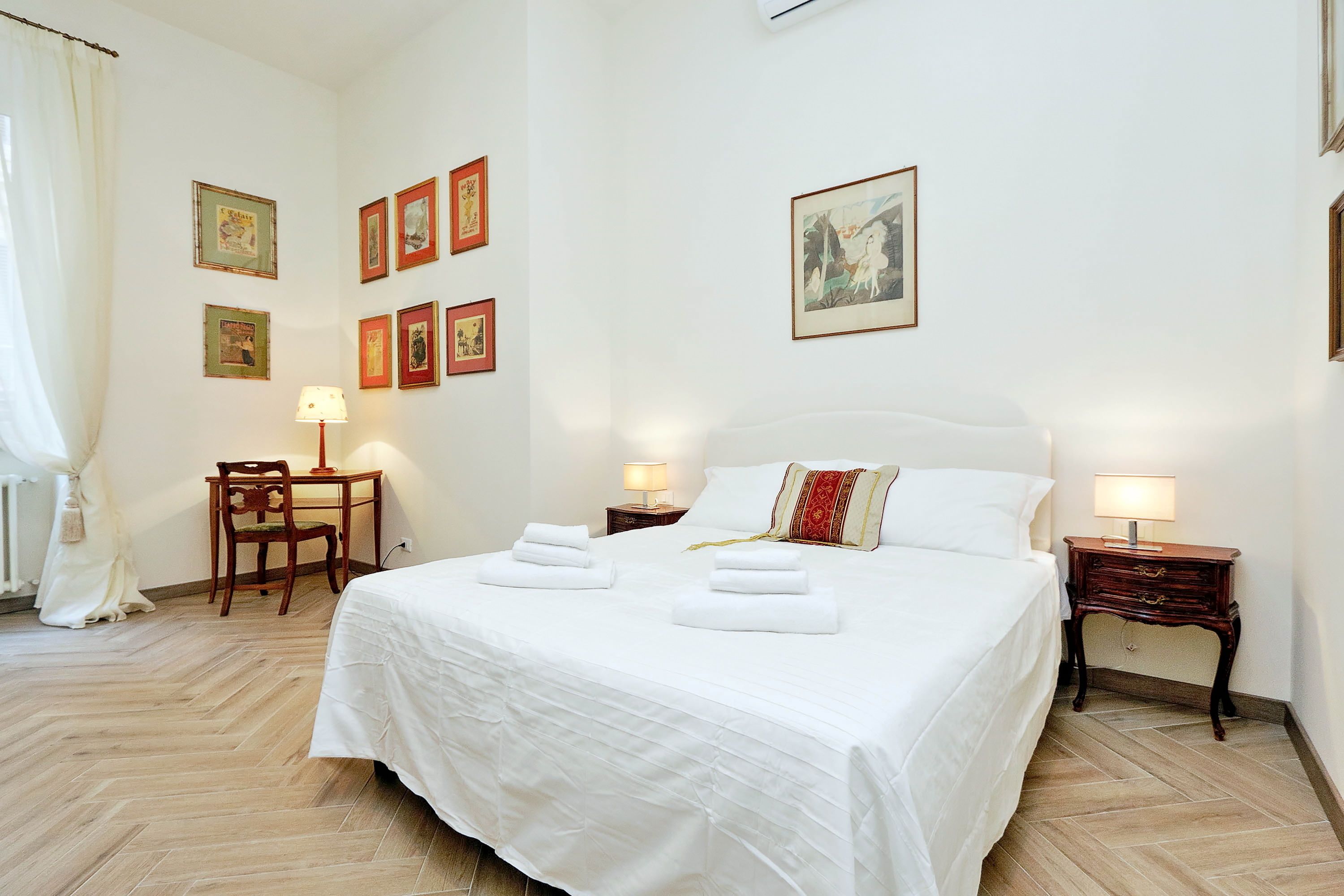 A neatly made bed with white linens and two towels, a wooden chair, a small table with a lamp, and several framed pictures on the wall.